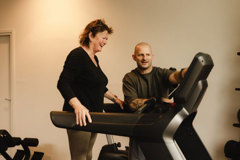 Oudere deelnemer lacht met haar personal trainer bij de loopband